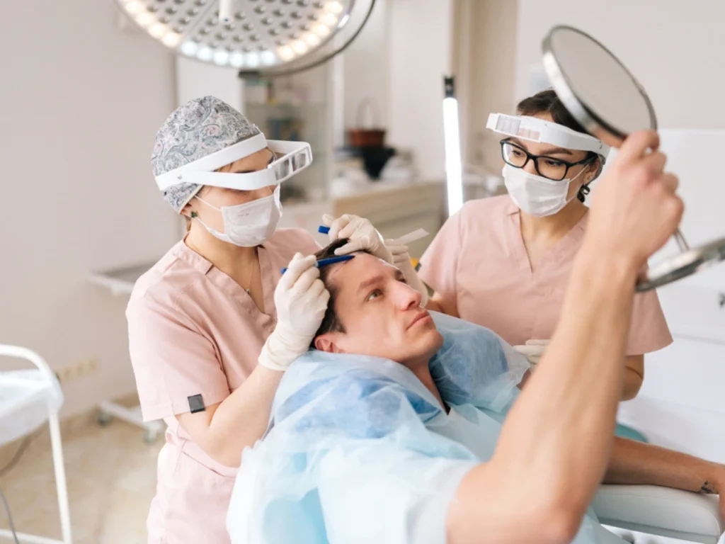 A patient examines his reflection while medical professionals prepare for Male Pattern Baldness Treatment.