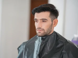 Man with a stylish haircut wearing cape in salon showcasing natural look with Mens Hair Systems.